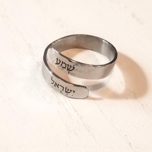 Puede incluir: Un anillo envolvente de color plateado con texto hebreo. El anillo tiene una textura martillada y presenta las palabras "Shema" e "Israel" grabadas en su superficie. El anillo se muestra sobre una superficie de madera blanca.