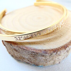 May include: A gold-colored cuff bracelet with engraved coordinates. The bracelet is resting on a light brown, circular wooden surface. The engraved text reads "32&deg;46'59.9"N 79&deg;..."