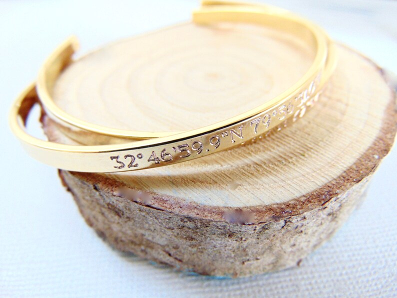 May include: A gold-colored cuff bracelet with engraved coordinates. The bracelet is resting on a light brown, circular wooden surface. The engraved text reads "32&deg;46'59.9"N 79&deg;..."