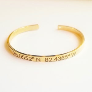 May include: A gold-colored cuff bracelet with engraved coordinates: 28.0552&deg; N 82.4385&deg; W. The open-ended bracelet is made of a shiny, polished metal. The bracelet is a simple, elegant design.