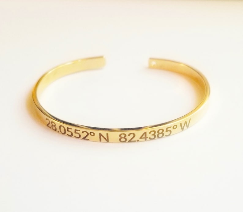 May include: A gold-colored cuff bracelet with engraved coordinates: 28.0552&deg; N 82.4385&deg; W. The open-ended bracelet is made of a shiny, polished metal. The bracelet is a simple, elegant design.
