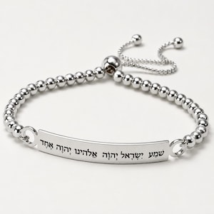 May include: A silver-toned adjustable bracelet featuring a central curved metal bar engraved with the Hebrew text "שְׁמַע יִשְׂרָאֵל יְהוָה אֱלֹהֵינוּ יְהוָה אֶחָד". The bracelet is constructed from a series of small, polished spherical beads and includes a sliding clasp mechanism.