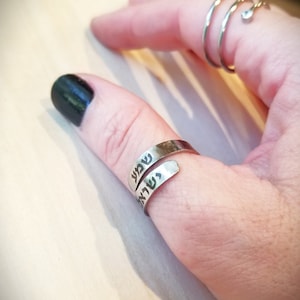 May include: A silver-toned wrap ring featuring the Hebrew text "Shema Yisrael" engraved in black lettering. The ring is worn on the thumb, while a separate silver-toned spiral ring with a small clear stone is worn on the index finger.