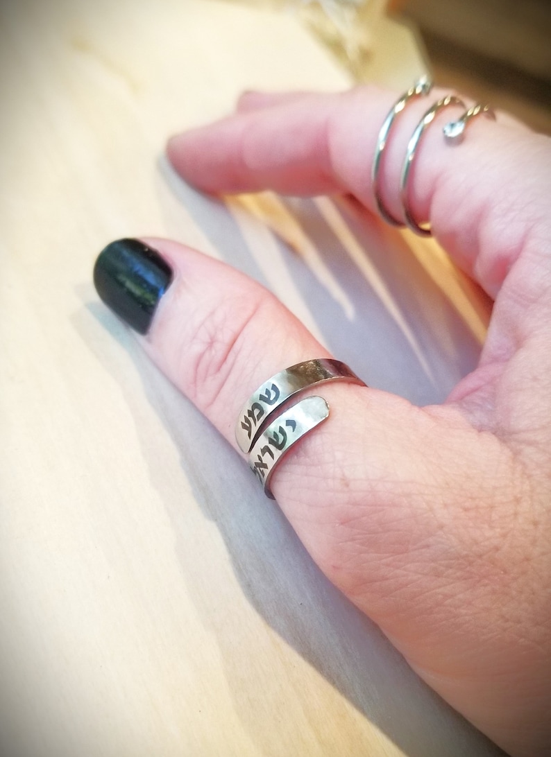 May include: A silver-toned wrap ring featuring the Hebrew text "Shema Yisrael" engraved in black lettering. The ring is worn on the thumb, while a separate silver-toned spiral ring with a small clear stone is worn on the index finger.