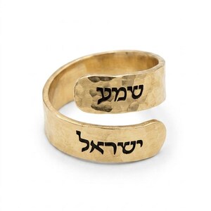 May include: A gold-toned bypass ring featuring a hammered texture on the metal surface. The ring displays black Hebrew text engraved on both ends of the open band, with the word 'שמע' on the upper section and 'ישראל' on the lower section.