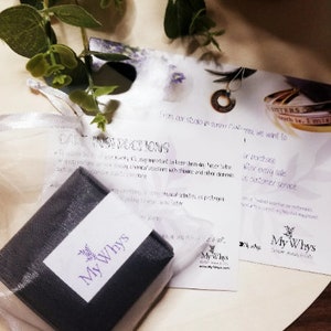 May include: A dark gray jewelry box with a white label featuring the purple My Whys logo, accompanied by a white care instruction card and a branded information card, both displaying the My Whys logo and text, alongside a green leafy sprig.