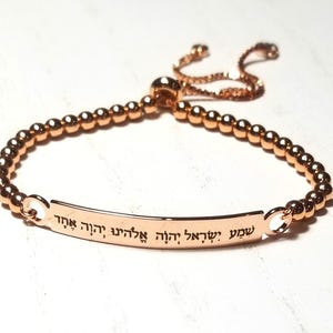 Può includere: Bracciale regolabile in oro rosa con catena di perline e barra rettangolare. La barra presenta testo ebraico inciso. Il bracciale è esposto su una superficie bianca.
