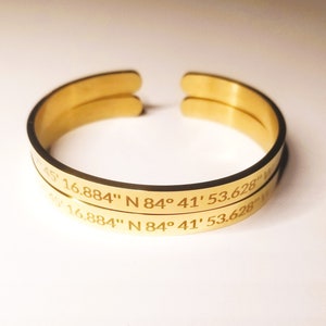 May include: Two gold-colored, open cuff bracelets. Each bracelet has engraved text: "45' 16.884" N 84&deg; 41' 53.628" W". The bracelets are stacked, with the text visible on the outer surface. The bracelets are made of a shiny, metallic material.