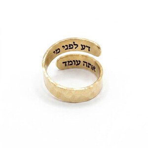May include: A gold-toned bypass ring with a hammered texture. The ring features black Hebrew text engraved on the inner surface, reading "דע לפני מי אתה עומד" across two overlapping bands, creating a continuous wrap-around design.