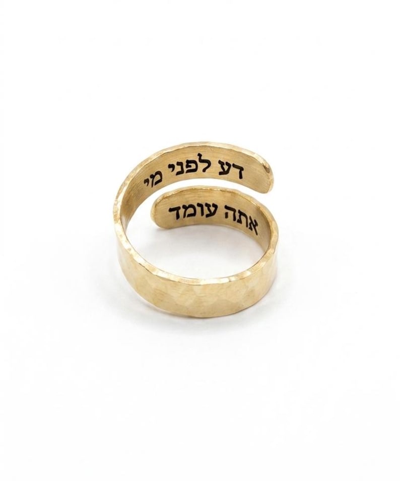 May include: A gold-toned bypass ring with a hammered texture. The ring features black Hebrew text engraved on the inner surface, reading "דע לפני מי אתה עומד" across two overlapping bands, creating a continuous wrap-around design.