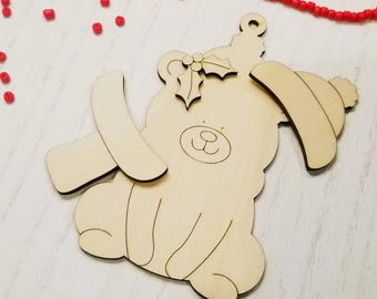 May include: A wooden Christmas ornament in the shape of a bear wearing a hat and scarf. The bear has a simple, cartoon-like design with etched details. A small loop is at the top for hanging. Red beads are scattered around the ornament.