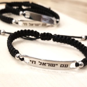 May include: Black braided bracelet with a silver rectangular tag engraved with Hebrew text. The text reads "סם אהבה" which translates to "Love Potion".