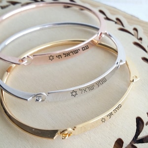 May include: Three gold bangle bracelets with engraved Hebrew text and a Star of David symbol. The bracelets are stacked on top of each other, with the gold bracelet on the bottom, followed by the silver bracelet, and the rose gold bracelet on top.