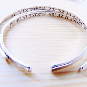 May include: Two silver-colored open cuff bracelets. The bracelets have engraved coordinates: 41&deg; 12.348"N 73&deg; 32' 19.4424"W and 29&deg; 45' 37.5372"N 95&deg; 22' 11.290"W. The bracelets are on a light wood surface.