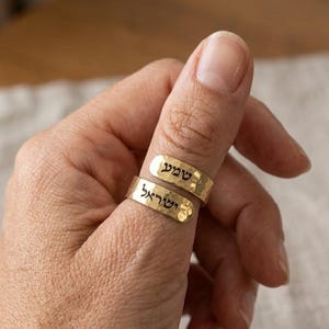 May include: Two gold-toned rings with a hammered texture, each featuring black Hebrew text engraved on the surface. The rings are stacked on a thumb, displaying the words "שמע" on the top band and "ישראל" on the bottom band.