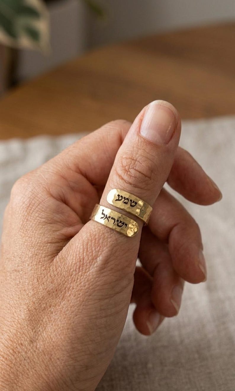 May include: Two gold-toned rings with a hammered texture, each featuring black Hebrew text engraved on the surface. The rings are stacked on a thumb, displaying the words "שמע" on the top band and "ישראל" on the bottom band.