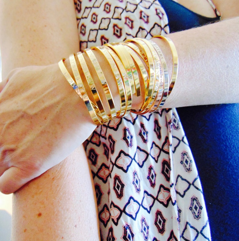 May include: A collection of gold-colored bangles stacked on a wrist. The bangles have a smooth, polished finish and some appear to have engraved text. The background includes a patterned top with a blue garment.