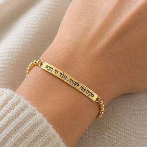 May include: A gold-toned beaded bracelet featuring a central rectangular bar engraved with black Hebrew text. The bracelet consists of small, uniform spherical beads strung together, creating a flexible band that encircles the wrist.