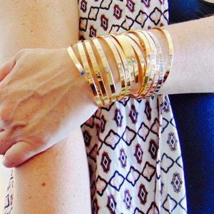 Puede incluir: Primer plano de un brazo con una pila de brazaletes de oro. Los brazaletes están apilados uno encima del otro y todos tienen el mismo ancho. La persona lleva una camisa con estampado blanco y negro.