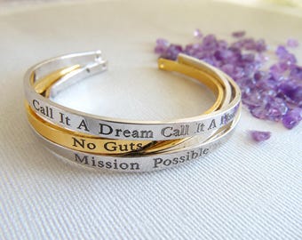May include: A set of three stacked metal cuff bracelets in silver and gold tones. The bracelets feature engraved text that reads Call It A Dream, Call It A Plan, No Guts, and Mission Possible. The cuffs have an open design and smooth finish.