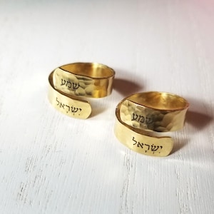 May include: Two gold-colored wrap rings with a hammered texture. Each ring features black Hebrew text engraved on the surface, displaying the words Shema Yisrael. The rings have an open, adjustable design with overlapping ends.