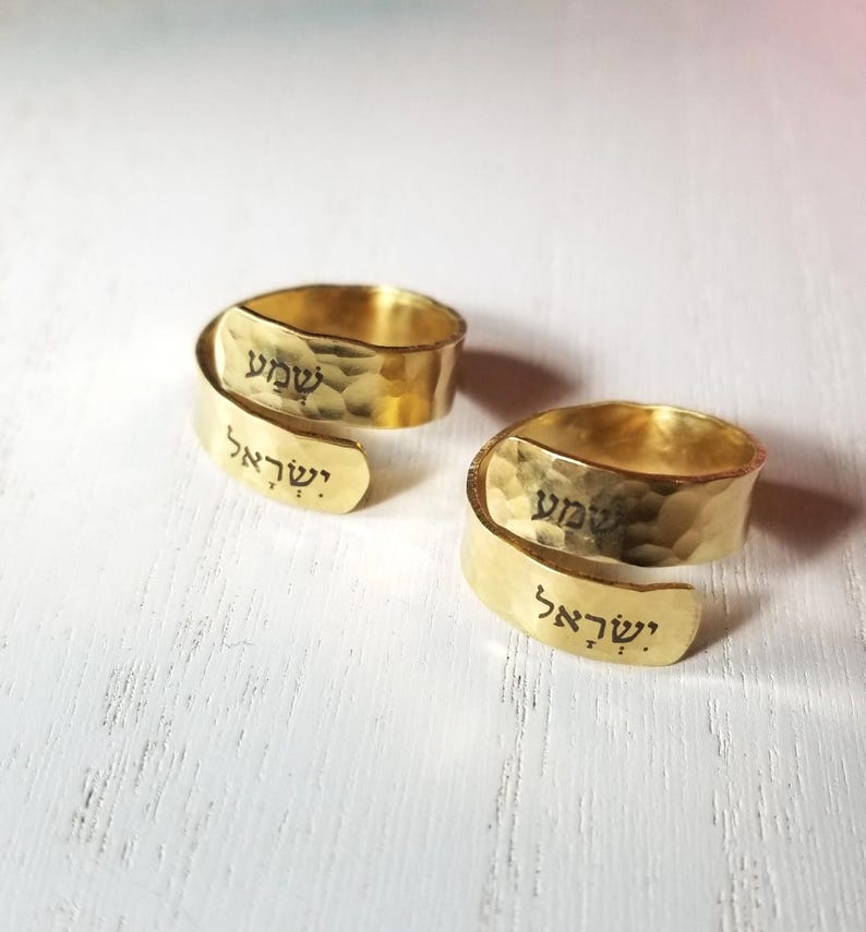 May include: Two gold-colored wrap rings with a hammered texture. Each ring features black Hebrew text engraved on the surface, displaying the words Shema Yisrael. The rings have an open, adjustable design with overlapping ends.