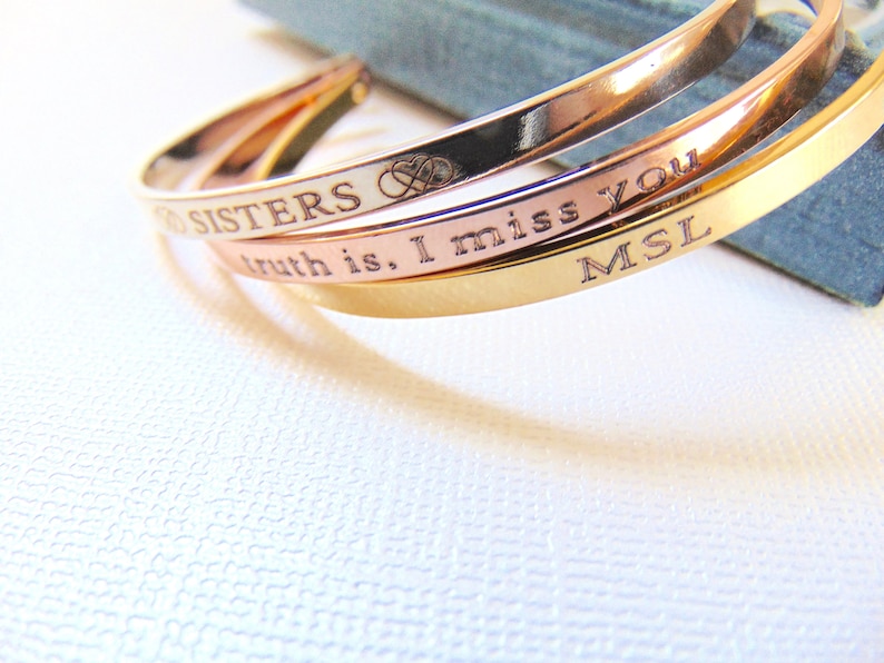 May include: Three gold, rose gold, and silver-colored engraved cuff bracelets. The bracelets have the phrases "Sisters", "truth is, I miss you", and initials "MSL" engraved on them. The bracelets are stacked together.