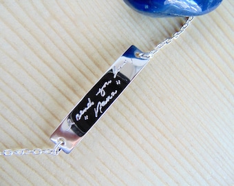 May include: A silver-toned rectangular metal bar pendant on a delicate chain, featuring the engraved cursive text "I love you Nana". A smooth, polished surface reflects light, and a portion of a dark blue stone is visible at the top.