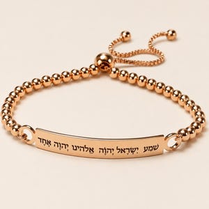 May include: A rose gold-toned beaded bracelet featuring a central curved metal bar engraved with the Hebrew text Shema Yisrael Adonai Eloheinu Adonai Echad. The bracelet includes a sliding adjustable closure with two small spherical end beads.