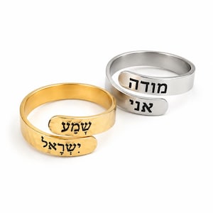 May include: Two adjustable wrap rings, one gold-toned and one silver-toned, featuring hammered metal bands. The gold ring is engraved with the Hebrew text "שמע ישראל" in black, while the silver ring is engraved with the Hebrew text "מודה אני" in black.
