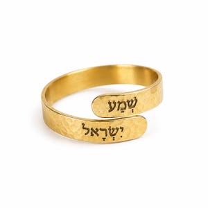 May include: A gold-colored bypass ring with a hammered texture. The ring features the Hebrew text "שמע" on the upper overlapping end and "ישראל" on the lower overlapping end. The metal has a polished finish and a wrap-around design.