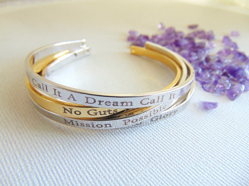 May include: Three open cuff bracelets in silver and gold tones. The silver bracelets have engraved text that reads "Call It A Dream", "No Guts", and "Mission". The gold bracelet is smooth. Amethyst crystals are scattered in the background.