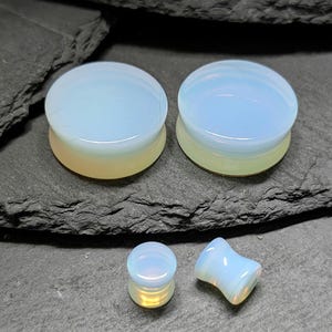 May include: Two large and two small circular ear plugs made of smooth, translucent opal-like material. The plugs feature a pale, milky white surface with subtle iridescent blue and yellow undertones, showcasing a polished finish and a concave shape.