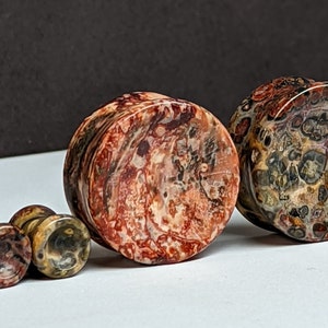 May include: Set of three stone ear gauges in various sizes. The gauges are made of a red, brown, and white patterned stone.