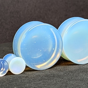 May include: Several opalescent ear plugs of varying sizes are arranged on a dark surface. The plugs have a light blue and white color scheme, with a rounded, smooth shape. The largest plugs are approximately 1 inch in diameter.