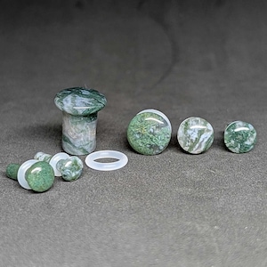May include: A collection of green and white stone plugs and a mushroom-shaped plug. The plugs vary in size and shape, with a mushroom-shaped plug and several round plugs. A white rubber ring is also included. The stone has a natural, marbled appearance.