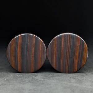 May include: Two round, wooden plugs with a striped pattern in shades of brown, red, and black. The plugs have a smooth, polished surface and are displayed against a dark background. These plugs are likely for ear piercings.