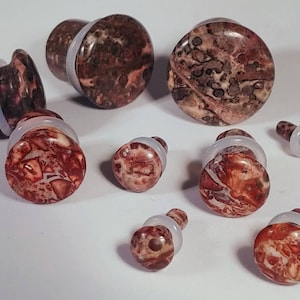 May include: A set of eight stone ear gauges in various sizes. The gauges are made of a red, brown, and white marbled stone. The gauges are all round and have a smooth finish.