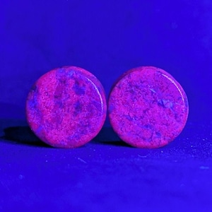 Puede incluir: Dos tapones redondos, rosas y morados con un patrón moteado. Los tapones están colocados sobre un fondo azul oscuro. Los tapones parecen estar hechos de piedra o un material similar.