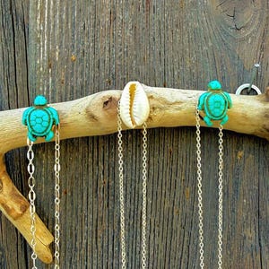 May include: A wall-mounted driftwood display rack featuring five hooks. The hooks alternate between white cowrie shells and turquoise-colored turtle figurines. Each hook supports a thin silver-toned metal chain hanging vertically downward.