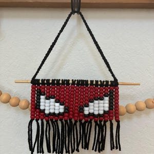 May include: A wall hanging made of red, white, and black plastic beads strung on black cord. The beads form two white eye-like shapes against a red background. It is suspended from a wooden dowel with a black cord loop and features black fringe.