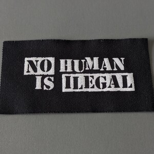 May include: A black rectangular fabric patch with the words "NO HUMAN IS ILLEGAL" printed in white, block letters. The patch has a textured appearance and is likely made of woven material.