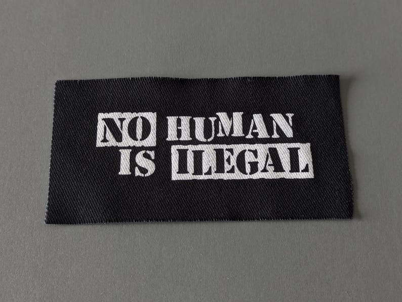 May include: A black rectangular fabric patch with the words "NO HUMAN IS ILLEGAL" printed in white, block letters. The patch has a textured appearance and is likely made of woven material.