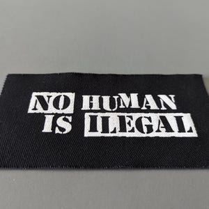 May include: A black fabric patch with the words "NO HUMAN IS ILLEGAL" printed in white, block letters. The patch has a rectangular shape with slightly angled edges and is set against a gray background.