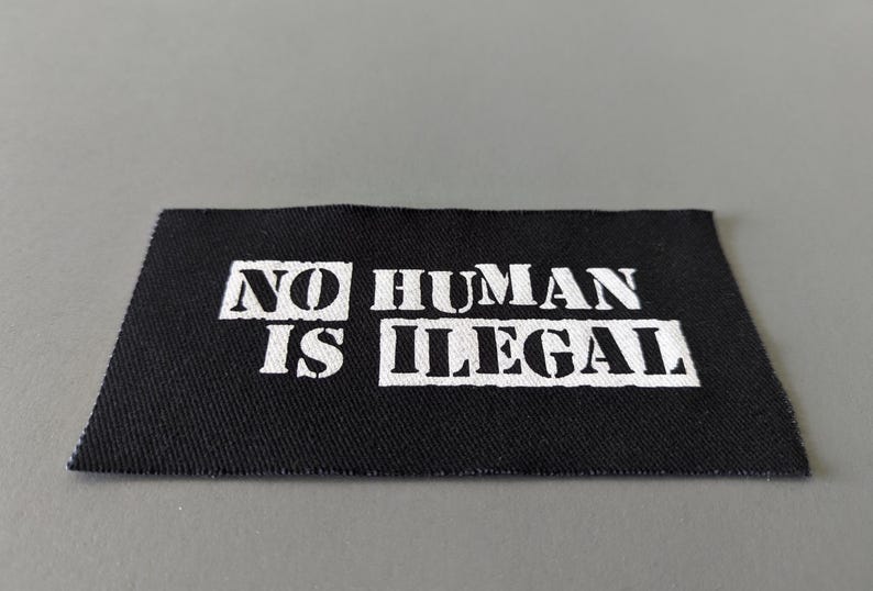 May include: A black fabric patch with the words "NO HUMAN IS ILLEGAL" printed in white, block letters. The patch has a rectangular shape with slightly angled edges and is set against a gray background.