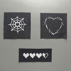 Puede incluir: Tres parches de tela negra con diseños serigrafiados en blanco. Un parche cuadrado presenta una telaraña con un corazón en el centro, otro parche cuadrado muestra un corazón de alambre de espino y un parche rectangular muestra cuatro iconos de corazones pixelados.