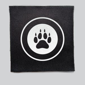 May include: Black square patch with a white paw print inside a white circle. The patch has a textured appearance, and the edges are frayed. The paw print is a symbol of animal love and care.