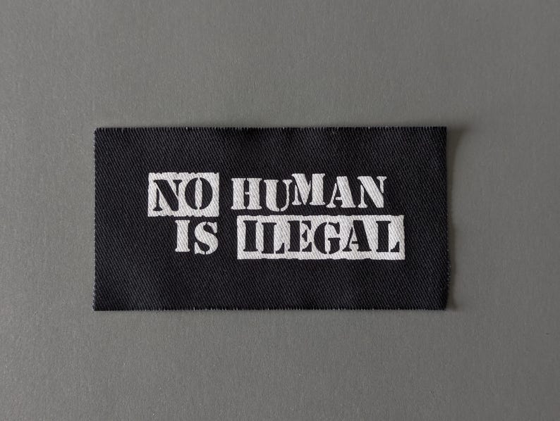 May include: A black rectangular patch with white text. The text reads 'NO HUMAN IS ILLEGAL' in a bold, sans-serif font. The patch has a slightly frayed edge, and the background is a solid gray color.