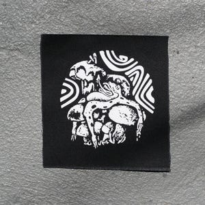 May include: Black square patch with a white graphic design. The design features a creature with tentacles and spots, surrounded by mushrooms and swirling patterns. The patch is likely for clothing or accessories.