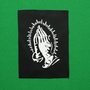 May include: A black rectangular patch with a white depiction of praying hands, encircled by a halo of radiating lines. The patch is set against a green background, providing a strong contrast.